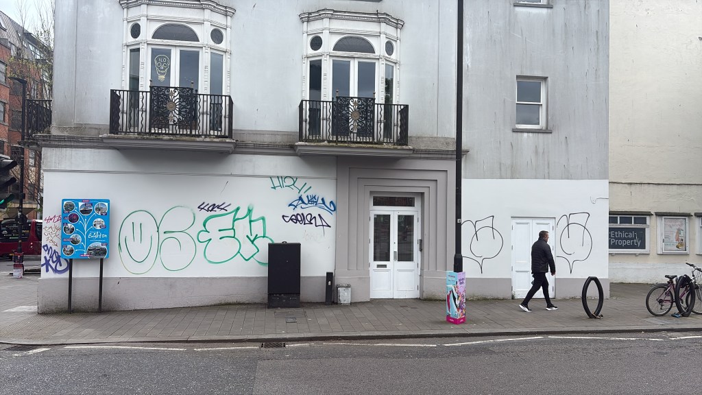 The north wall of The Queen's Head opposite Brighton Station: white render covered in graffiti tags, with a doorway in the middle and a passer-by walking past.